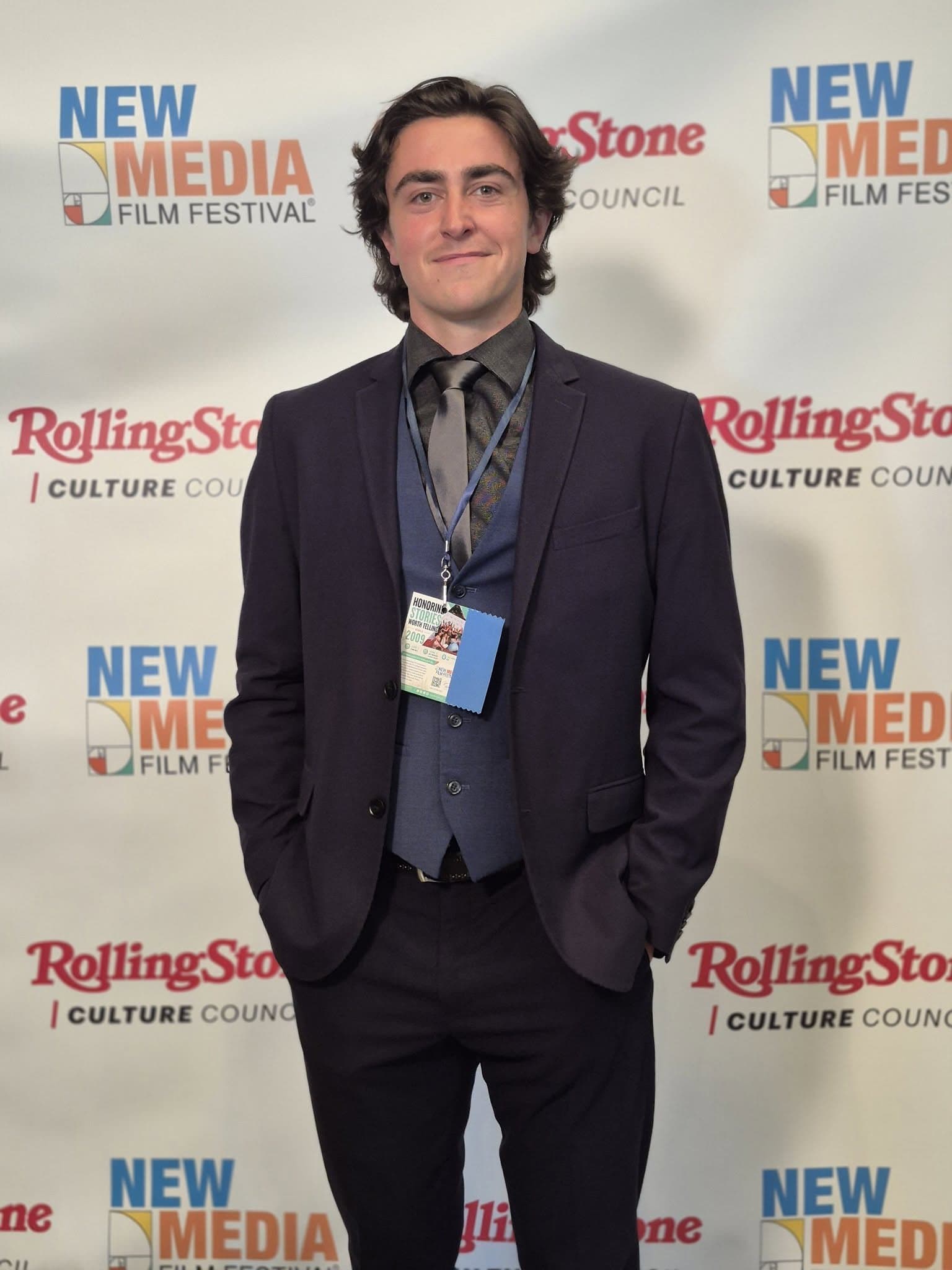 Owen Logan, wearing a suit and a festival badge, stands in front of a New Media Film Festival step-and-repeat backdrop.