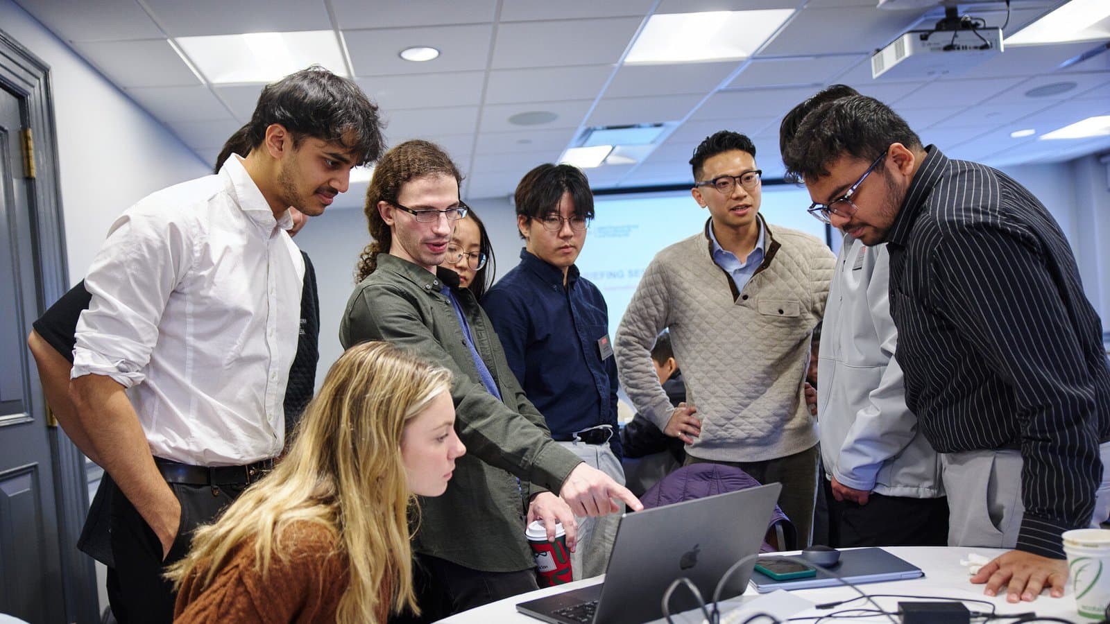 A group of graduate students gathers around a laptop on a conference table while one student points at the screen.